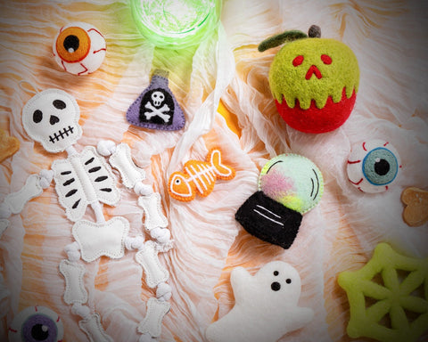 A collection of handmade felt Halloween-themed accessories and decorations arranged on a textured white and orange surface. The set includes a white felt skeleton with bones, a green and red pumpkin, a white ghost, a yellow spider web, several animal eyes, a purple skull and crossbones patch, an orange fish bone, and a glowing green cup.